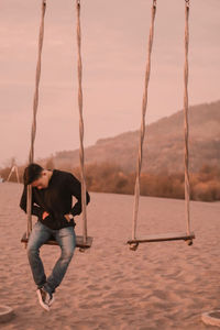 Man on swing at beach against sky during sunset