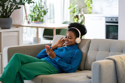 Young relaxed african woman in headphones holding phone listening favorite music or podcast at home