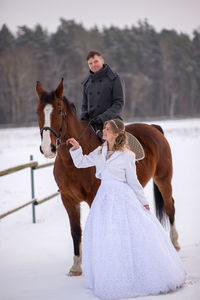 Portrait of smiling groom sitting on horse by bride on snow covered field