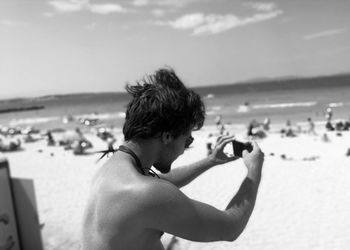 Shirtless man photographing at beach against sky