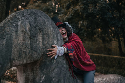 Side view of man cosplaying mad hatter wearing top hat sitting on statue against trees