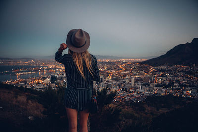 Rear view of woman by illuminated cityscape against clear sky during dusk