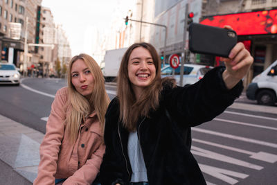 Cheerful female friends standing on street in madrid and taking self shot on smartphone during city stroll