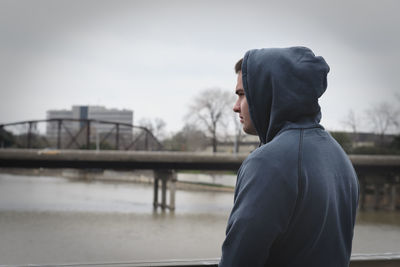 Young man wearing hooded jacket while standing by river against sky