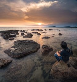 Rear view of man sitting on rock at beach during sunset