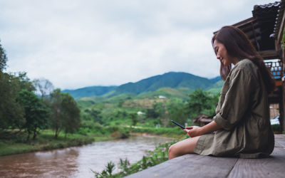 Young woman using phone while sitting on mountain against sky