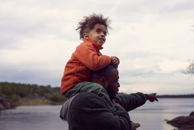 Father pointing at sea while carrying son on shoulders against sky