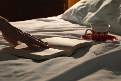 Cropped image of hand with book and coffee cup on bed at home