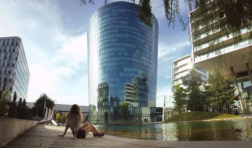 Woman sitting in front of infinity pool by modern building in city against sky