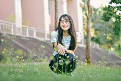 Portrait of young woman crouching on grass against building at park