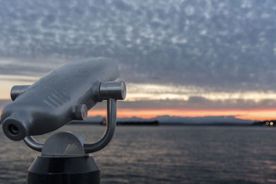 Coin-operated binoculars by sea against cloudy sky during sunset