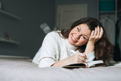 Young adult woman forty years body positive in white sweater writes notes in planner on bed at home