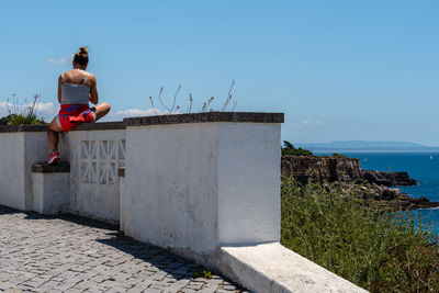 Rear view of man sitting on retaining wall against clear sky