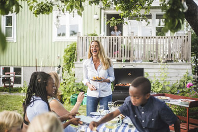 Happy mature woman standing with grilled food plate while friends sitting at table in yard during weekend