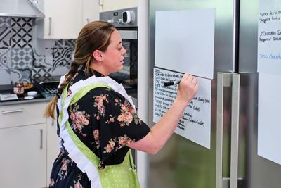 Side view of female in apron standing and writing notes on paper hanging on fridge while cooking in light kitchen in daytime