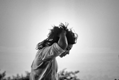 Side view of young woman with hand in hair standing against clear sky