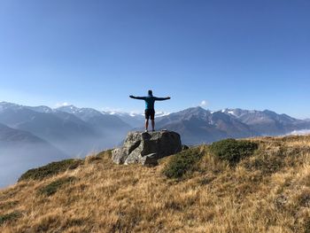 Rear view of man with arms outstretched while standing on rock at mountain against sky