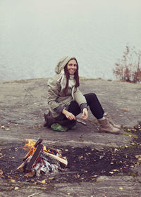 Full length of woman holding stick by campfire at lakeshore