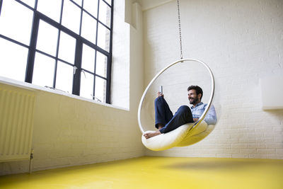 Smiling man sitting on swing in his loft taking selfie with smartphone