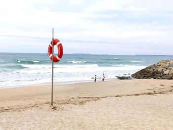 Life belt on pole at beach with people carrying surfboards against sky