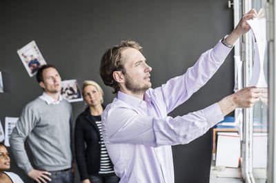 Mid adult businessman sticking paper on window with colleagues looking at him in office