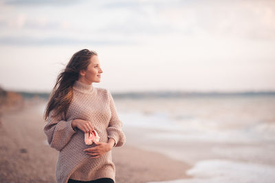 Smiling pregnant woman wear knit cardigan hold baby shoes posing over sea background close up.