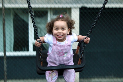 Smiling girl sitting on swing at playground against fence