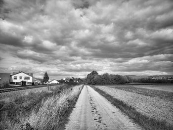 Dirt road amidst field against cloudy sky