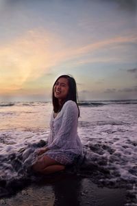 Portrait of young woman sitting at beach against sky during sunset