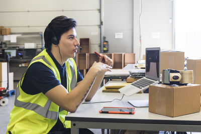 Side view of confident young male customer service representative talking through headset while sitting at desk in wareh