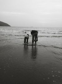 Rear view of father and son rolling pants while standing on shore