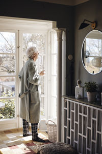 Senior man in bathrobe holding coffee cup while looking outside window at home