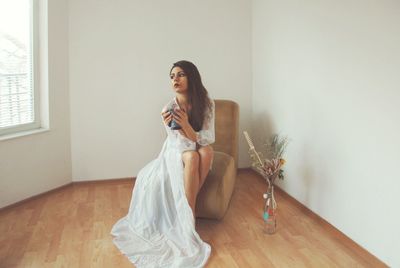 Young fashion model sitting with drink on chair against wall