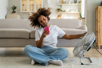 African american girl sitting on floor in living room near electric fan and using smartphone