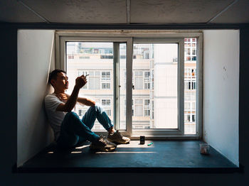 Young man smoking cigarette while sitting by window at home