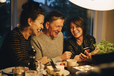 Smiling senior woman sharing smart phone with friends while sitting at dining table during dinner party