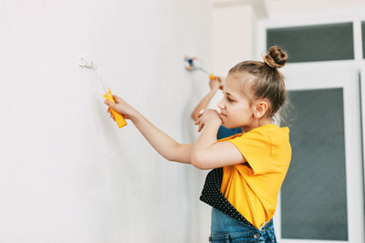 A girl in a denim overalls and a yellow t-shirt helps to paint the walls in an apartment white.