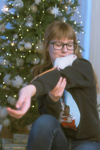 Girl holding rat while sitting against christmas tree