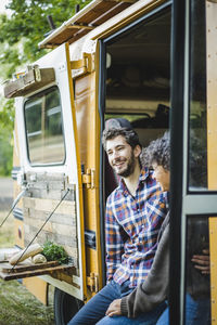 Happy young multi-ethnic couple sitting at caravan doorway during camping in forest