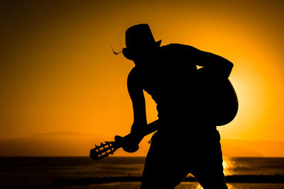 Silhouette man playing guitar at beach against orange sky