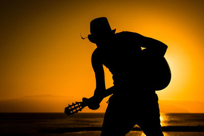 Silhouette man holding camera at beach against sky during sunset