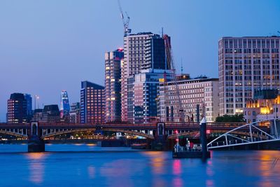 Bridge over river against illuminated buildings in city at dusk