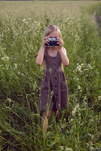 Little girl child in a checkered dress stands with a retro camera and photographs in summer field