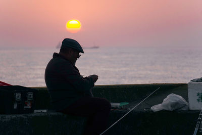 Man sitting by sea against sky during sunset