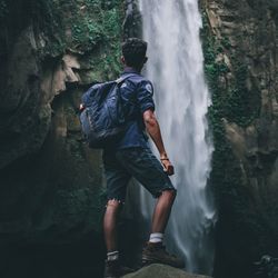 Rear view of man standing on rock against waterfall in forest