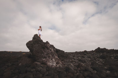 Low angle view of young man standing on rock against cloudy sky