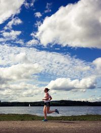 Side view of mature woman running on field by lake against cloudy sky