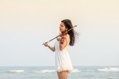 Portrait of young woman playing violin at beach against clear sky