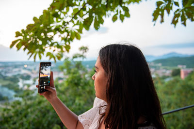Side view photo of young woman taking photo of landscape view with mobile phone