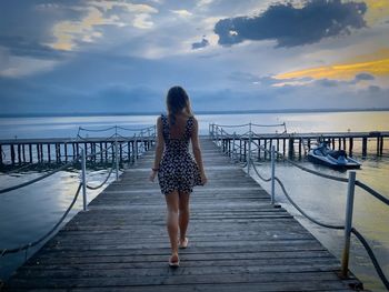 Woman in summer dress walking down a wooden pontoon towards the sea at sunset